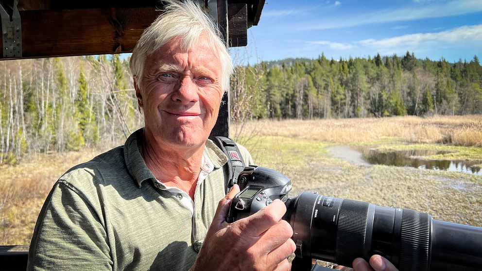 Vithårig man med en stor kamera ler mot kameran framför en bakgrund av skog och vass.