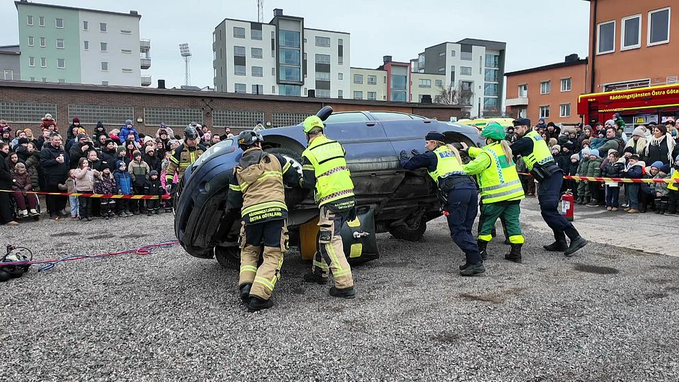 Brandmän och räddningspersonal välter en bil på en grusplan inför en publik som står bakom avspärrningar. Flera åskådare har samlats för att titta på demonstrationen, och moderna lägenhetsbyggnader syns i bakgrunden.