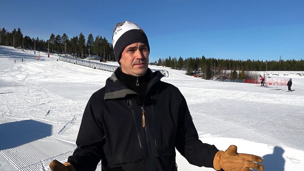 Person i skidjacka och mössa står på snötäckt skidanläggning med klarblå himmel.