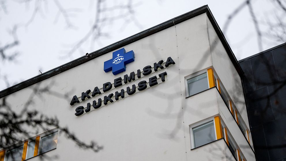 Klocka på Akademiska sjukhuset med tydlig logotyp och namnskylt på fasaden, omgiven av trädgrenar.