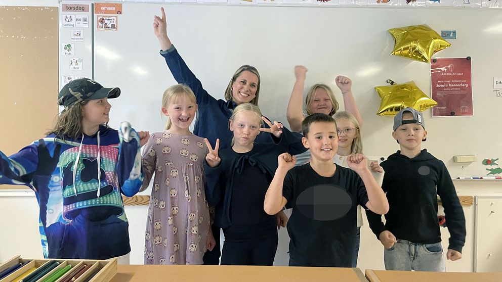Grupp av barn som poserar framför en whiteboard, glada och applåderande. Några har händerna upp i luften och visar tecken på seger. Bakom dem hänger två gyllene stjärnor dekorationer. Klassen verkar vara i en skolmiljö.