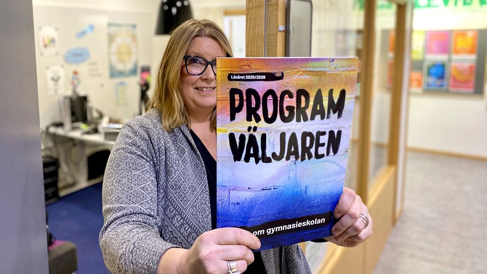 En person står i en korridor och håller upp en färgglad broschyr med texten ”Program Väljaren” som verkar handla om gymnasieskolan. I bakgrunden syns ett kontor och en korridor med färgglad dekor.