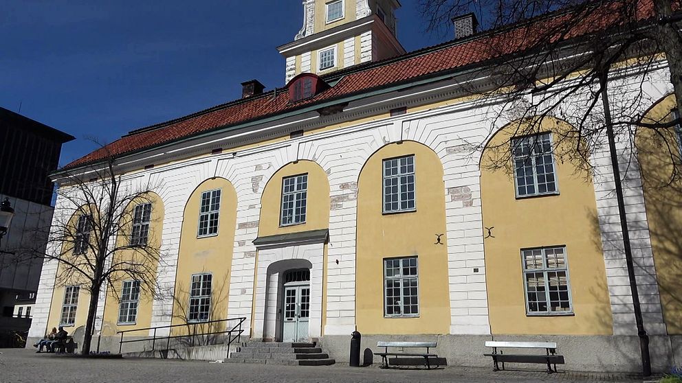 Gul och vit byggnad med flera fönster, blå himmel och några träd i förgrunden. Fasaden är skadad.