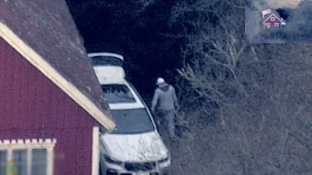 En man i grå träningskläder står vid en vit bil som är parkerad vid skogskanten. I bakgrunden syns ett rödaktigt hus med vita fönster. Området är omgiven av tät vegetation.