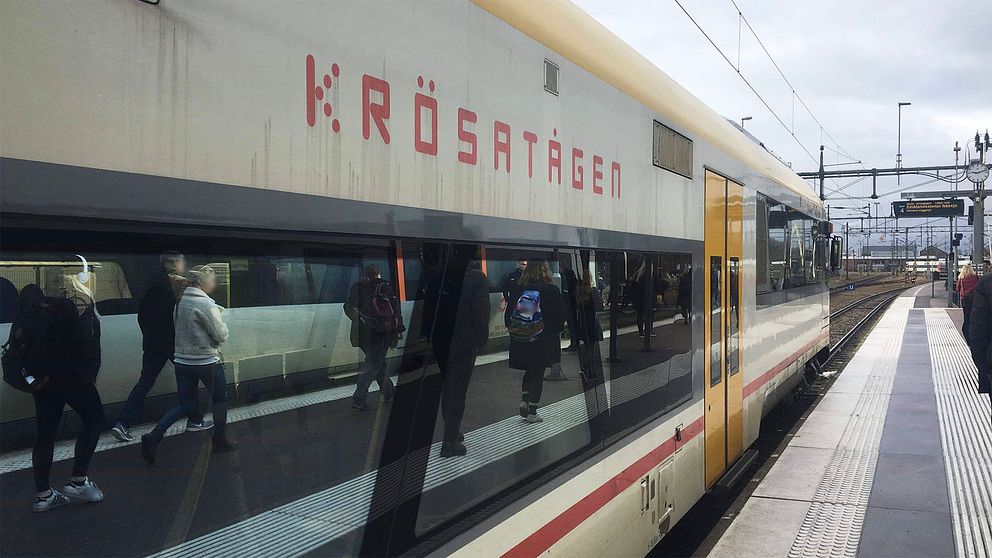 Ett Krösatåg står still på perrongen.  Texten på tåget är röd, dörrarna är gula och i tågets fönster speglas resenärer på perrongen.