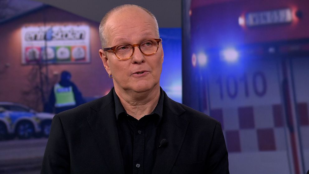En person i mörk klädsel syns i förgrunden. I bakgrunden finns en scen med en polisbil, en polis och en byggnad med skyltar samt en brandbil med nummer 1010.