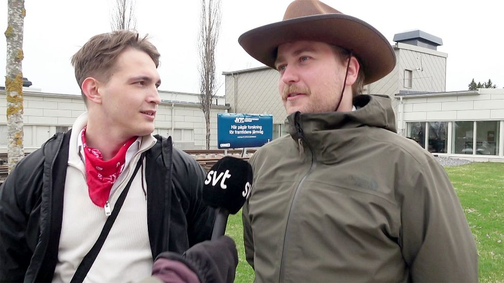 Två personer intervjuas utanför en byggnad. En av dem har en cowboyhatt på huvudet.