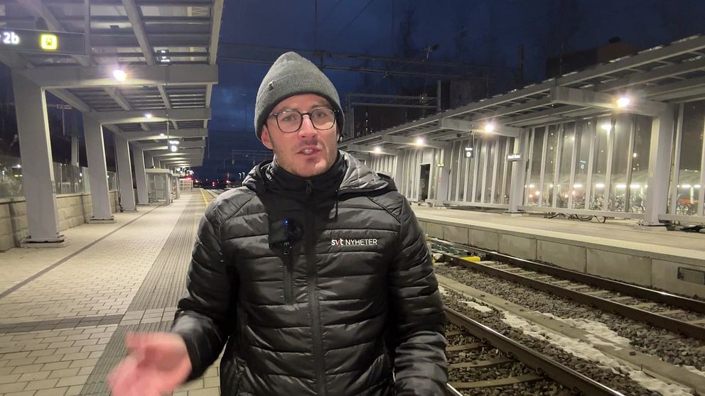 En person står på en tågstation under kvällstid, iförd en svart jacka och en mössa. Stationen har en modern arkitektur med lyktor som skapar en belyst miljö. Tågrälsen syns i bakgrunden tillsammans med stationens plattform.