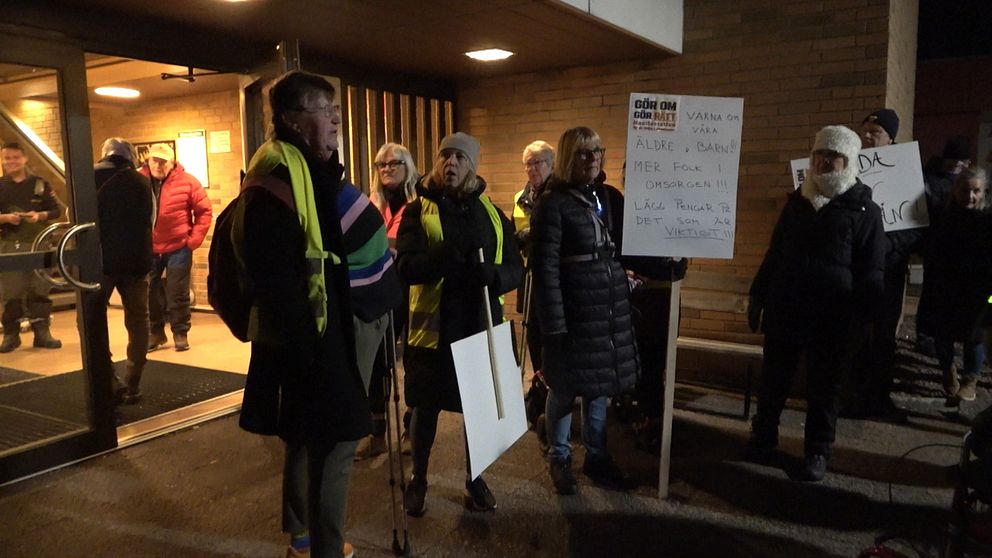 Människor samlas utanför en byggnad på kvällen, några står i grupp med skyltar. En person bär en färgglad sjal, medan andra har reflexvästar. En stor skylt med text är placerad framför gruppen. Bakgrunden visar andra människor som observerar händelsen.