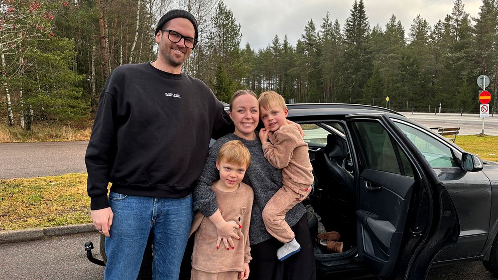 En familj står utomhus vid en bil, omgivna av träd och en vägskylt i bakgrunden. Två barn står framför en vuxen, som håller ett av barnen i famnen, medan det andra barnet står bredvid. De är klädda i avslappnade kläder och ler mot kameran.