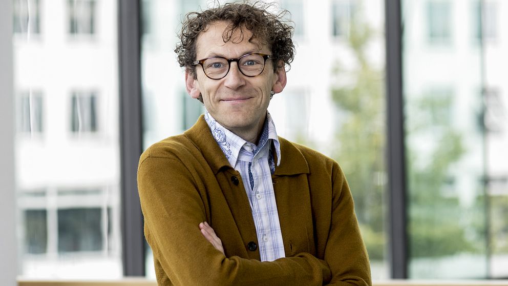 En person, Åke Ingerman, med korslagda armar står i ett ljust rum med stora fönster som släpper in naturligt ljus. Personen har på sig en brun kofta över en mönstrad skjorta. I bakgrunden syns oskarpa kontorsmiljöer.