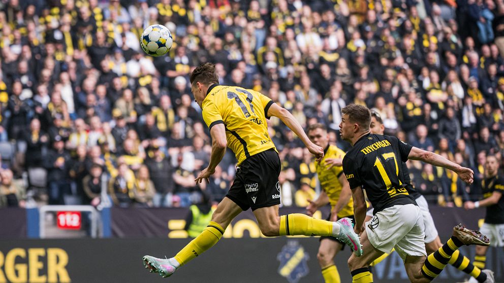 Fotbollsspelare i gult gör ett nickmål under match mot lag i svart framför stor publik.