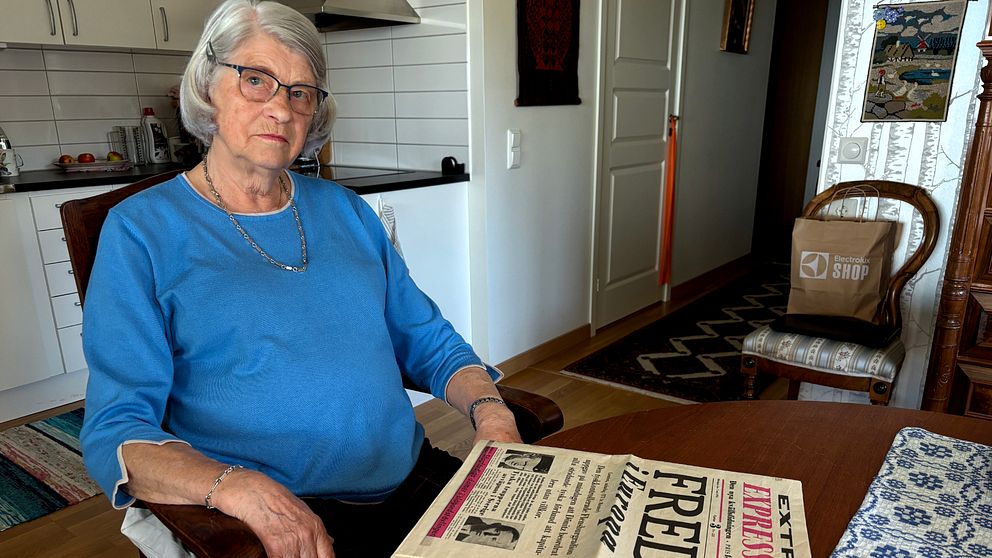 Äldre person i blå tröja sitter vid köksbord och läser en tidning med rubriken ”Fred”.