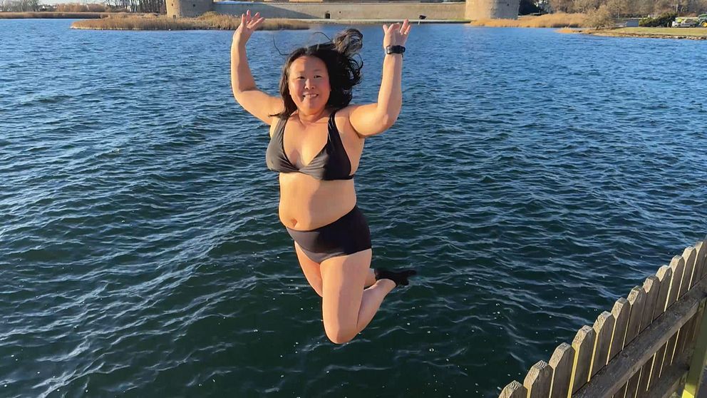En person är i luften på väg att hoppa i vattnet från en brygga, iförd en bikini. Vattnet är klart och reflekterar solljuset, och i bakgrunden syns Kalmar slott. Det är en solig dag, vilket skapar en livlig och energisk atmosfär.