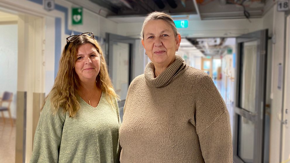 Två personer står i en sjukhuskorridor, klädda i komfortabla tröjor. Korridoren har flera dörrar och släpljus som ger en lugn atmosfär. Bakgrunden är suddig men visar att det är en vårdmiljö.