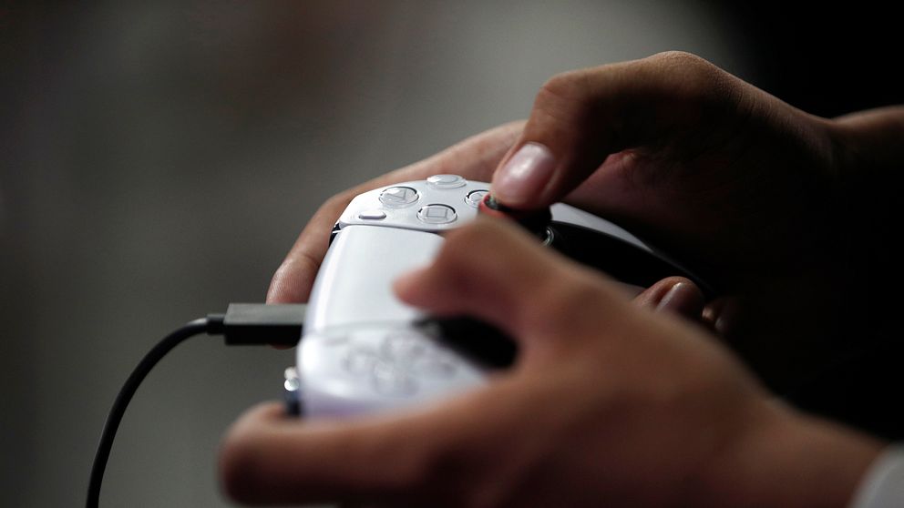 Händer som håller en vit spelkontroll, troligtvis en PlayStation 5-kontroll, i en mörk bakgrund.