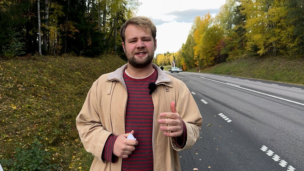 En person står vid vägrenen på en landsväg omgiven av träd med höstfärger. I bakgrunden syns flera fordon på vägen under en molnig himmel. Personen gestikulerar med ena handen, vilket kan indikera en pågående diskussion eller presentation.