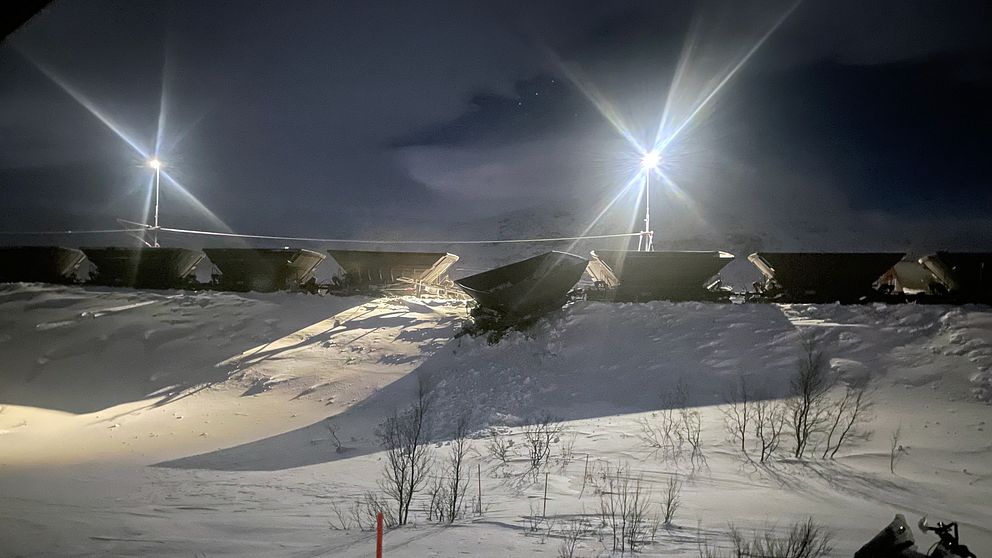 strålkastare över malmvagnar på vall, snötäckt mark. En vagn hänger ner längs vallen