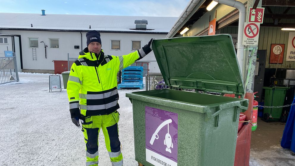 En person i en reflexjacka står vid en öppen grön avfallscontainer märkta med texten ”Fiskeredskap” på en vintrig plats med snö. Bakgrunden visar en del av en byggnad och andra containrar i närheten.