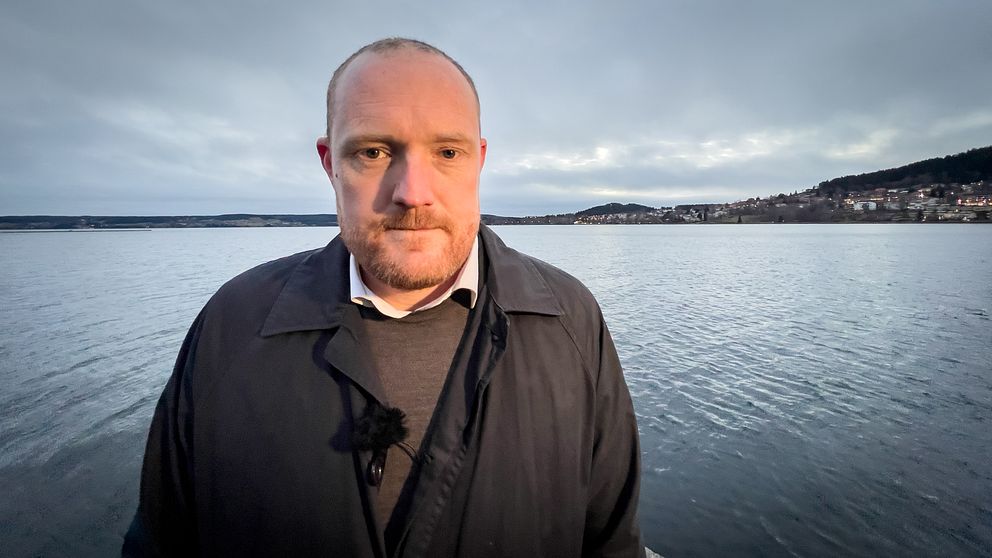 En person står vid vattnet med en sjö och en strandlinje i bakgrunden. Himlen är mulen och skapar en dämpad atmosfär. Personen är klädd i en mörk kappa och har ett mikrofonfäste på bröstet.