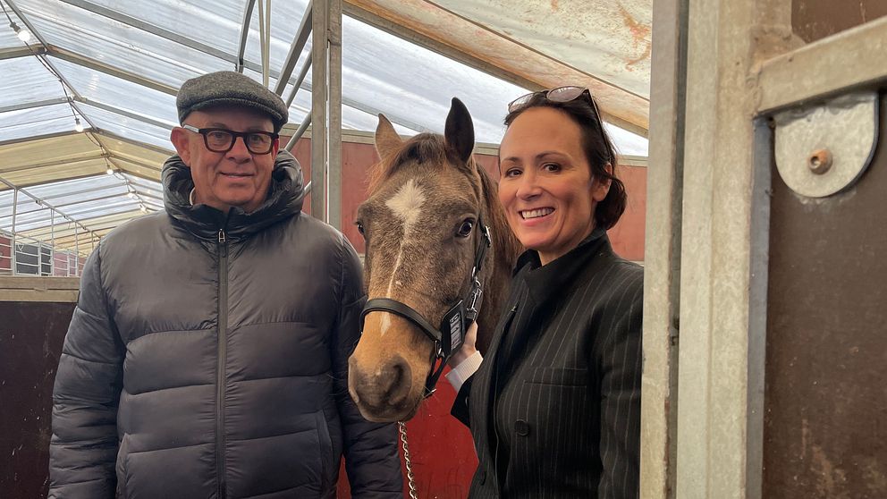 En person i en svart pufferjacka står till vänster om en häst med brunt och vitt huvud, medan en person i en svart kostym står till höger om hästen. De befinner sig i en stallmiljö med synliga stallväggar och tak.