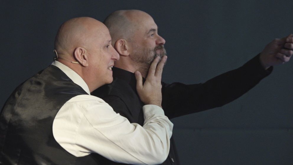Två män står tätt intill varandra på scenen. Den ena håller handen i ansiktet på den andre.