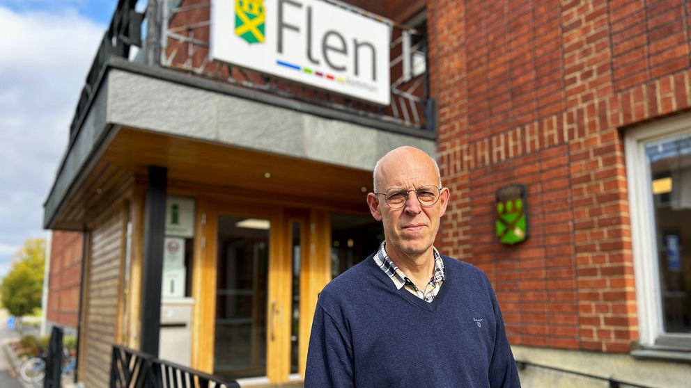 En person står framför entrén till ett byggnad med en skylt som visar namnet ”Flen”. Byggnaden har trä- och tegelarkitektur, och det finns en grön emblem på väggen. Det är en klar dag med lite moln på himlen.