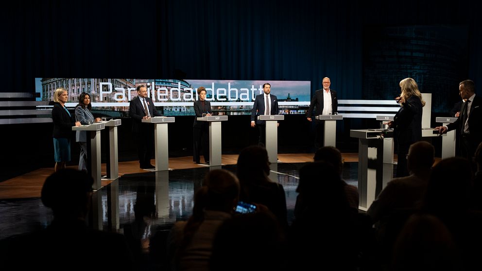Partiledardebatt på scenen, partiledare står bakom varsitt podium, publik i förgrunden.