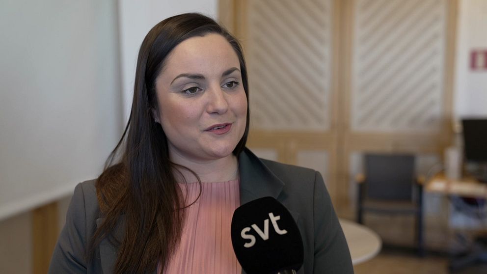 Kvinna i rosa tröja med SVT-mikrofon, stol och bord i bakgrunden.