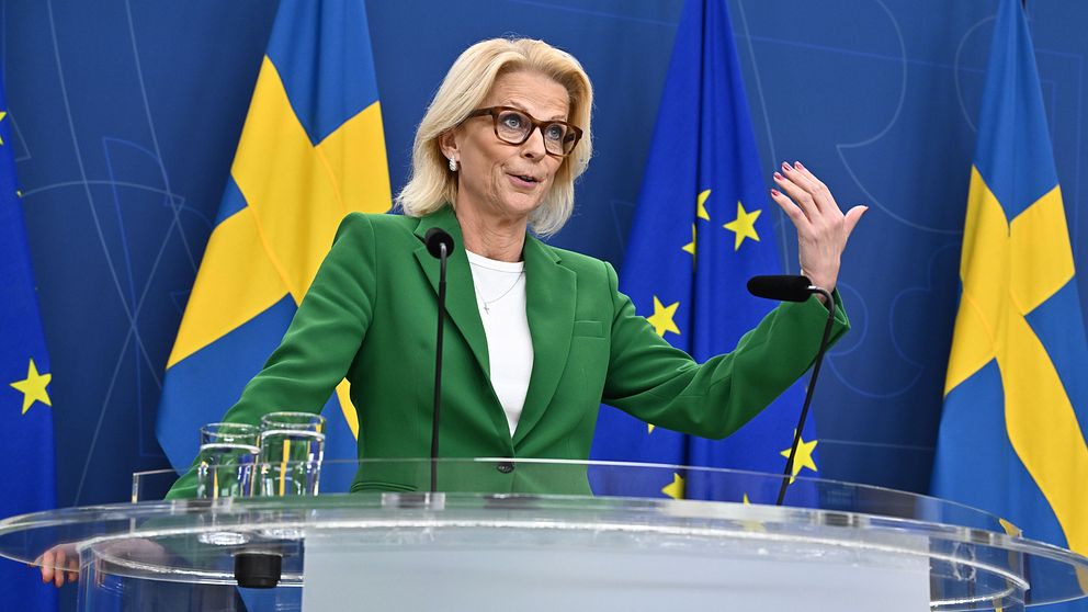 Finansminister Elisabeth Svantesson i grön kavaj står vid en talarpodium med glas vatten och en mikrofon, omgiven av svenska och europeiska flaggor. Hon gestikulerar med ena handen. Texten på podiumet visar ”Regeringen”.