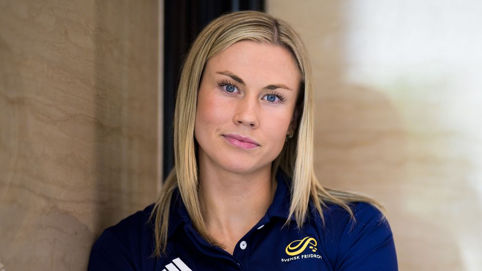 Person i mörkblå sporttröja står lutad mot en ljus vägg, armar i kors.