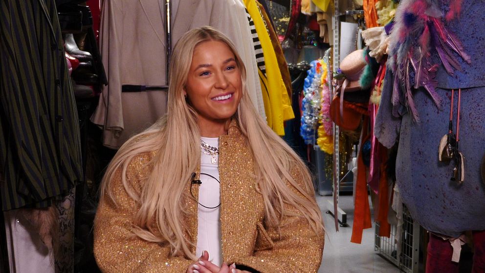 En person med långt blont hår sitter omgiven av färgglada kläder och accessoarer i en garderob. Personen bär en glittrig kappa över en vit topp. I bakgrunden syns olika klädställningar fyllda med plagg i olika stilar och färger.