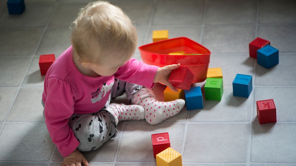 Ett litet barn i en rosa tröja sitter på golvet och leker med färgglada byggklossar, medan en röd behållare står öppet bredvid. Klossarna är spridda omkring barnet, och några klossar har bokstäver eller symboler på dem.