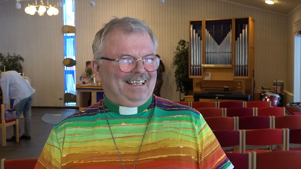 En kyrka med orgel och rader av röda stolar i bakgrunden. En man i regnbågströja ler.