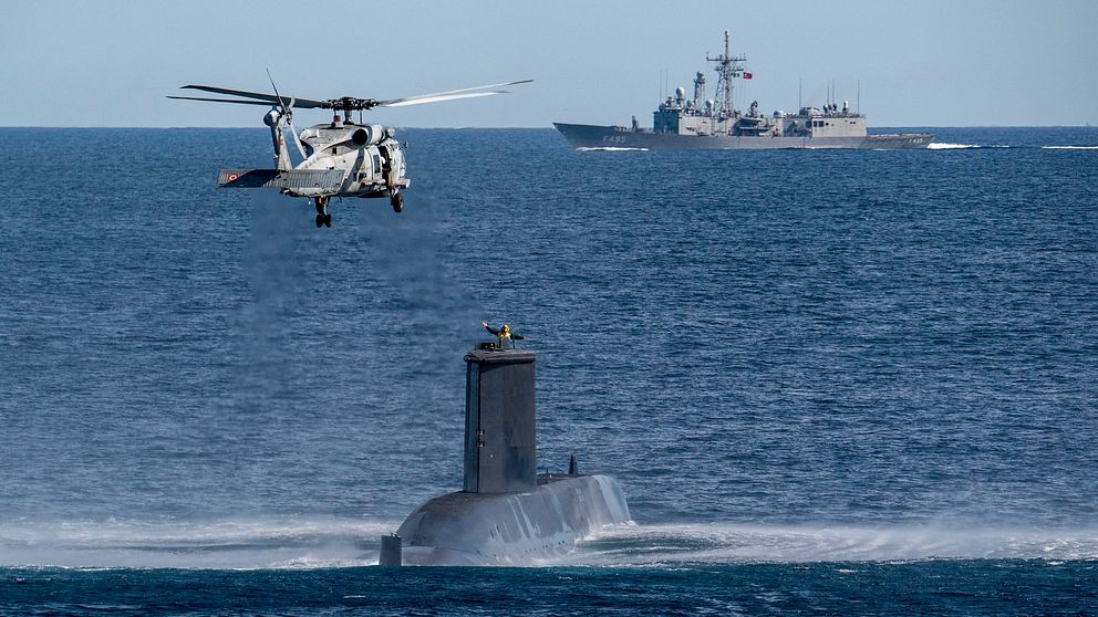 Helikopter ovanför ubåt på havet, krigsfartyg i bakgrunden.
