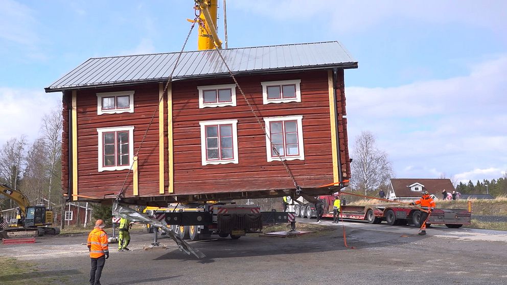 Hembygdsgården i Hackås lyfts av en stor kranbil till en väntande trailer. Byggarbetare i varselkläder övervakar arbetet. Huset är timrat och rött med vita foder runt fönstren.