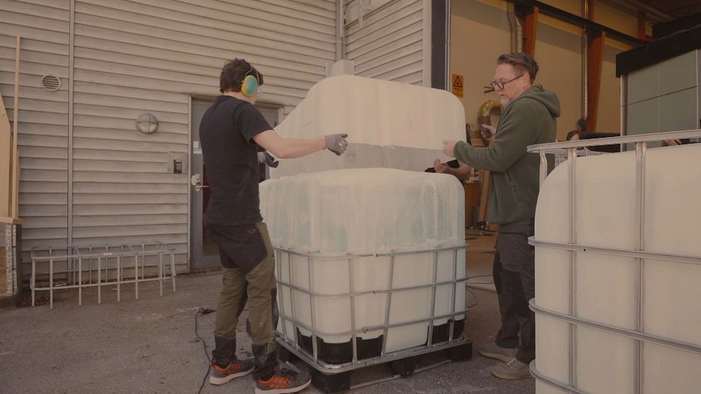 Två personer hanterar en stor  plastbehållare med is i en industrilokal.