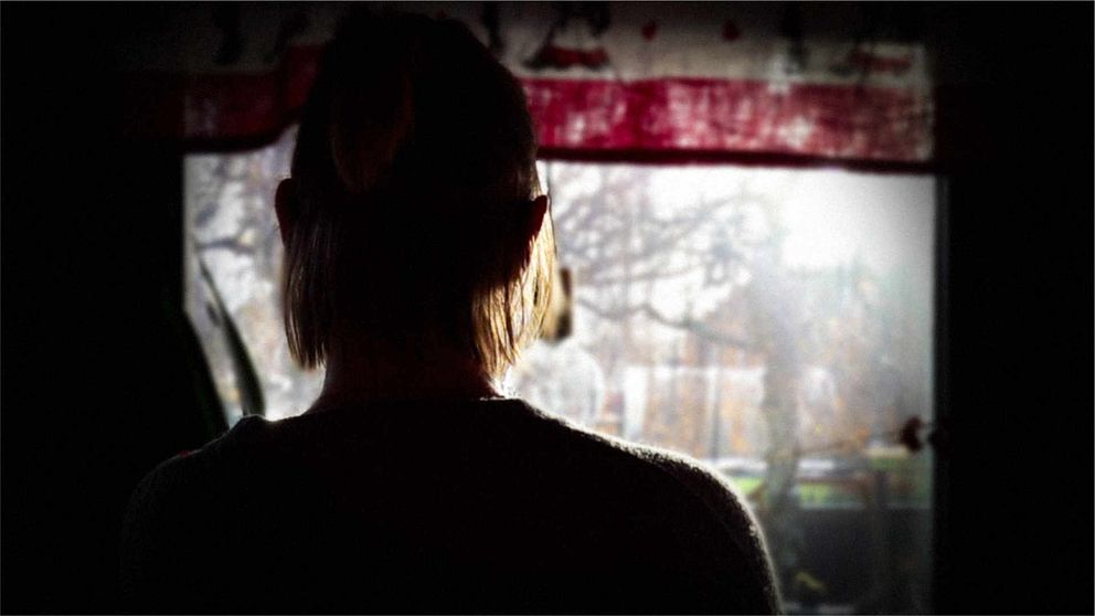 En person med kort hår står vänd mot ett fönster, där dagsljus strömmar in. I förgrunden syns silhuetten av personen, medan utsidan är suddig och belyses av det naturliga ljuset. Mörka toner dominerar bilden, vilket skapar en stämning av eftertänksamhet.