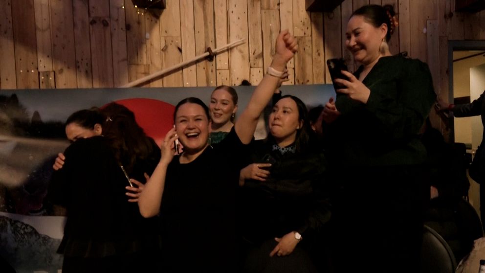 En grupp människor firar med höjda händer, stående nära en trävägg.