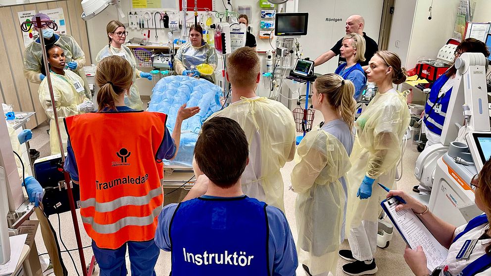 En grupp sjukvårdspersonal i skyddskläder samlas i ett sjukhusrum, ledda av en person i orange väst med texten ”Traumaledare” och en annan i en blå väst med texten ”Instruktör”. De verkar delta i en medicinsk träning eller simulering med medicinsk utrustning och en säng i mitten av rummet.