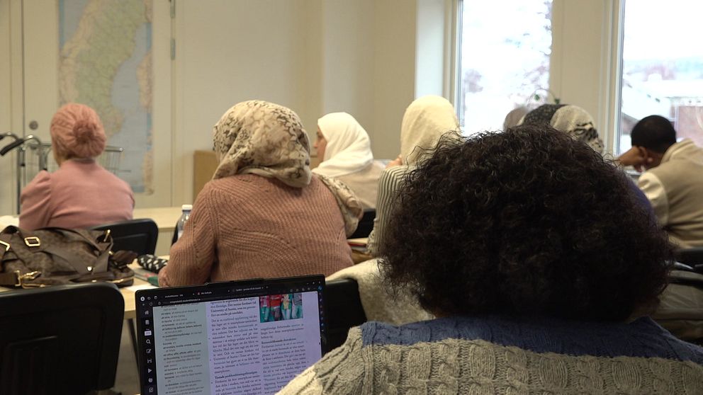 Ett klassrum, med tre rader. Det sitter flera personer i klassrummet och bilden är tagen bakifrån personerna. Längst fram i klassrummet hänger en karta över Sverige.