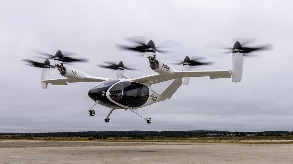 En svartvit elektrisk drönare med flera rotorer som lyfter från en landningsplats. Drönaren har en aerodynamisk design med tydliga propellrar i rörelse, vilket indikerar att den är i flygning. I bakgrunden syns en grönskande och öppen landskap under en övercast himmel.