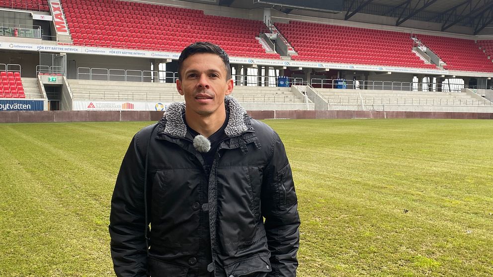 En person i mörk jacka står på en gräsplan inuti en idrottsarena med tomma röda sittplatser i bakgrunden. Arenans läktare och bås är tydligt synliga.