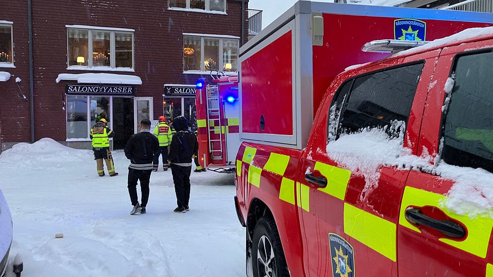 Brandmän i vinterkläder arbetar på en snötäckt gata utanför en restaurang, där en brandbil med blåljus står parkerad. Vid entrén till restaurangen syns flera personer, troligen räddningstjänstpersonal och eventuell personal från restaurangen, som diskuterar situationen. Snön ligger djup på marken och byggnaden är utrustad med stora fönster där ljus syns inifrån.