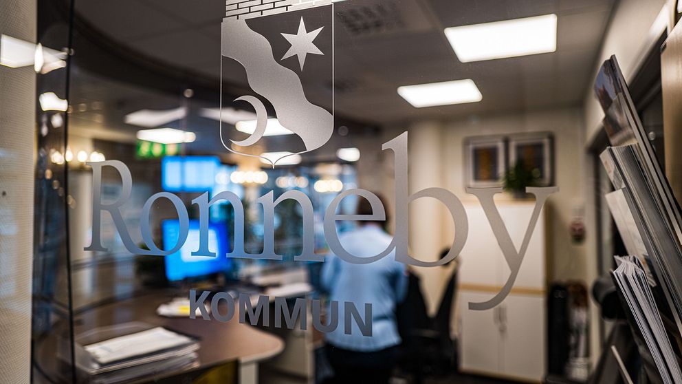 ALT-text: Skylt med texten ”Ronneby kommun” på en glasdörr, med suddig bakgrund som visar ett kontor. En person i blå skjorta syns delvis i bakgrunden.