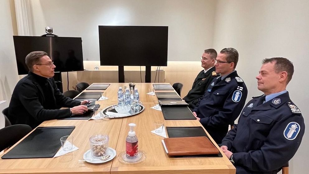 En konferensrumsscen med en träbord där en person i svart kläder sitter vid ena änden av bordet och arbetar på en laptop, medan tre uniformerade poliser sitter på andra sidan.