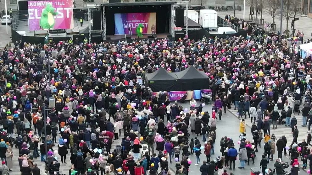 En stor folkmassa deltar i en festival, med en scen i bakgrunden där en artist uppträder. Många i publiken bär färgglada hattar och kläder, vilket skapar en festlig atmosfär. Scenens backdrop har texten ”Kom in till MelFest”, vilket antyder att evenemanget är en musikfestival.