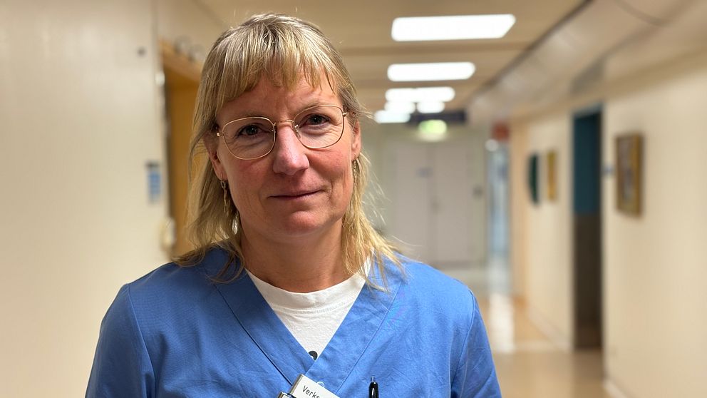 En sjuksköterska iklädd blå arbetskläder står i en lång korridor på ett sjukhus. Namnskylt och en penna syns i bröstfickan. Korridoren är ljus med vitt och blått som dominerande färger.