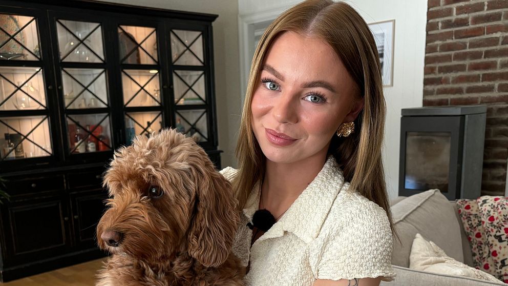 Kvinna med tatuerad arm sitter i ett vardagsrum och håller en brun hund i knät.