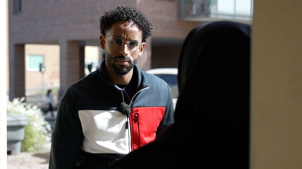 En person står utomhus och pratar med någon vid en ytterdörr i ett bostadsområde.